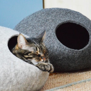 May include: Two gray felt cat beds, one with a cat inside. The cat beds are shaped like a rounded oval with a hole for the cat to enter. The cat bed on the left is light gray and the cat bed on the right is dark gray.