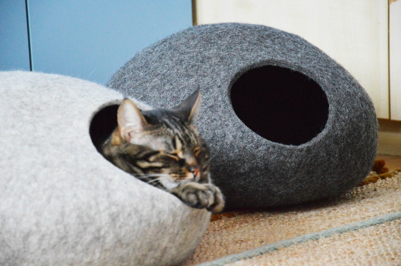 May include: Two gray felt cat beds, one with a cat inside. The cat beds are shaped like a rounded oval with a hole for the cat to enter. The cat bed on the left is light gray and the cat bed on the right is dark gray.