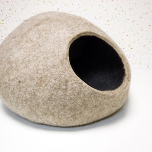 Pode incluir: Uma cama para gatos de feltro bege em forma de oval arredondado com um interior de feltro preto. A cama tem uma grande abertura de um lado.