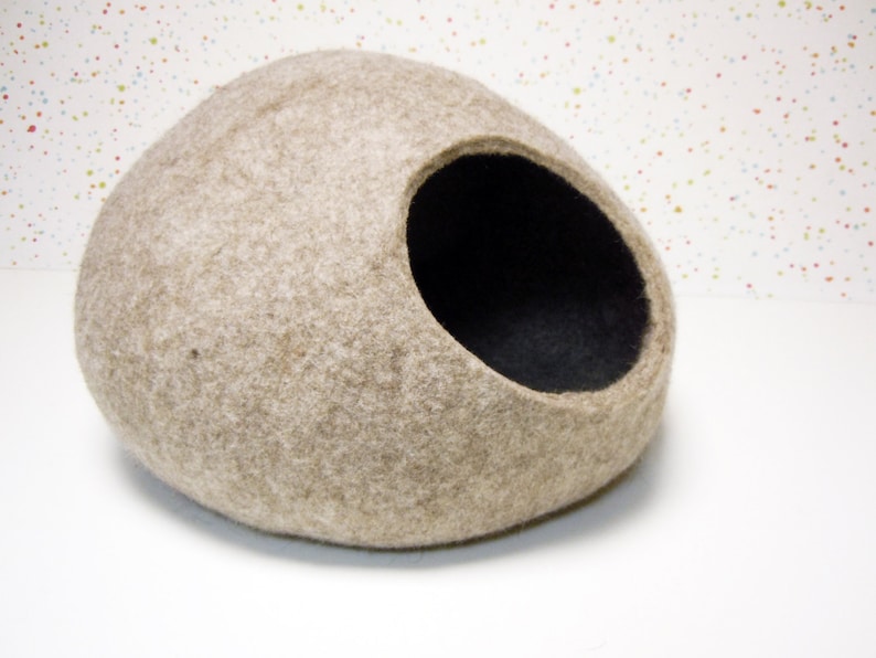 Pode incluir: Uma cama para gatos de feltro bege em forma de oval arredondado com um interior de feltro preto. A cama tem uma grande abertura de um lado.