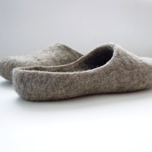 K&ouml;nnte beinhalten: Ein Paar graue Filzschuhe. Die Hausschuhe sind in einem ged&auml;mpften Grauton gehalten und haben ein einfaches Schlupfdesign. Das Filzmaterial wirkt weich und bequem, ideal f&uuml;r den Innenbereich. Die Hausschuhe werden auf einer wei&szlig;en Oberfl&auml;che gezeigt.