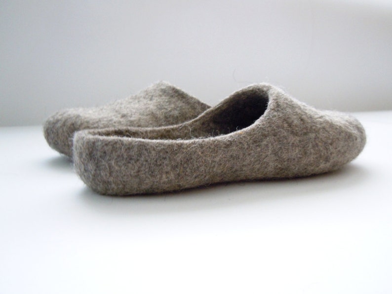 K&ouml;nnte beinhalten: Ein Paar graue Filzschuhe. Die Hausschuhe sind in einem ged&auml;mpften Grauton gehalten und haben ein einfaches Schlupfdesign. Das Filzmaterial wirkt weich und bequem, ideal f&uuml;r den Innenbereich. Die Hausschuhe werden auf einer wei&szlig;en Oberfl&auml;che gezeigt.