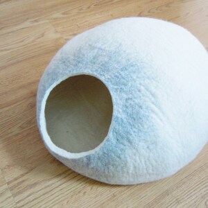 May include: A white and gray felt cat bed shaped like a dome with a large opening for entry. The bed is on a wooden floor.