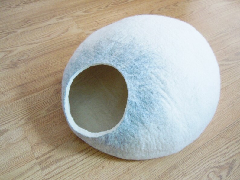 May include: A white and gray felt cat bed shaped like a dome with a large opening for entry. The bed is on a wooden floor.