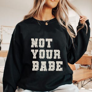 May include: Black sweatshirt with the text "NOT YOUR BABE" in a distressed, off-white font. The sweatshirt is a crew neck style and appears to be made of a soft material. The overall aesthetic is casual and comfortable.