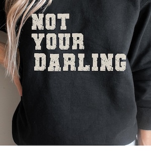 Könnte beinhalten: Schwarzes Sweatshirt mit dem Text "NOT YOUR DARLING" in einer verwitterten, cremefarbenen Schrift. Das Sweatshirt hat eine einfarbige Farbe und scheint aus einem weichen Material zu sein. Der Text ist mittig auf der Brust platziert.