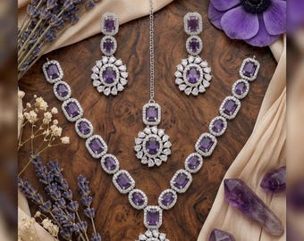 May include: A complete jewelry set featuring a necklace, earrings, and a headpiece. The set is adorned with purple rectangular and round gemstones, surrounded by smaller clear stones. The jewelry is displayed on a wooden surface with lavender and a purple flower.