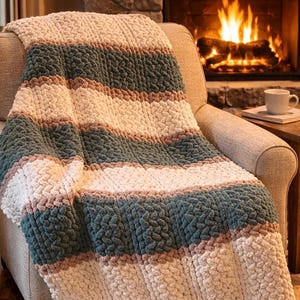 Può includere: Una coperta a righe nei toni crema, verde acqua e beige drappeggiata su una poltrona beige. La coperta a maglia grossa presenta spesse righe orizzontali. Un camino con fuoco acceso è visibile sullo sfondo, creando un'atmosfera calda e accogliente.