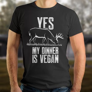 Może przedstawiać: Czarna koszulka z napisem "YES" nad grafiką jelenia i napisem "MY DINNER IS VEGAN" w kolorze białym. Jeleń jest przedstawiony podczas wypasu na trawie. Koszulka ma okrągły dekolt.