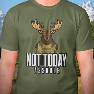 May include: An olive green short-sleeve t-shirt featuring a graphic of a brown moose wearing a tan tactical vest. Below the moose, the text "NOT TODAY ASSHOLE" is printed in bold white capital letters with a black outline on the front of the shirt.