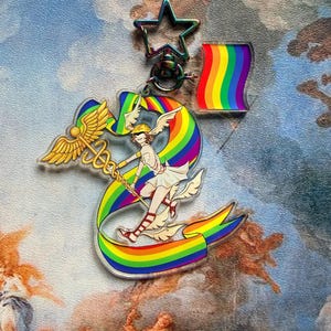 May include: An acrylic keyring featuring a winged figure holding a caduceus staff, surrounded by a rainbow ribbon. The charm includes a star-shaped clasp and a separate small square rainbow flag charm, both with an iridescent metallic finish.