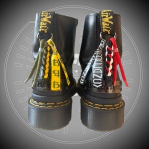 May include: A pair of black leather boots with thick soles and yellow stitching. The back of each boot features a black heel loop with yellow text, adorned with decorative hanging charms in olive, yellow, white, black, and red braided cords.