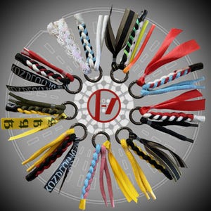 May include: A collection of twelve circular metal keychains, each featuring a unique arrangement of colourful fabric ribbons, braided cords, and shoelaces. Some ribbons display the text KOZDLOU, while others are solid, striped, or patterned.