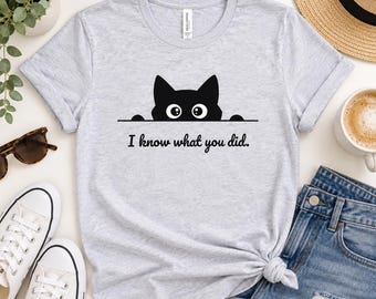 May include: A light gray short-sleeve t-shirt featuring a black graphic of a cat peeking over a horizontal line with the text "I know what you did." printed in black below the cat. The shirt has a crew neckline and a standard fit.