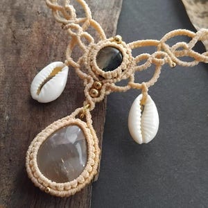 Könnte beinhalten: Eine beige Makramee-Halskette mit einem zentralen tropfenförmigen braunen Stein, einem kleineren runden dunklen Stein und zwei weißen Kaurimuscheln. Das komplexe Design aus geknoteter Kordel enthält kleine goldfarbene Perlen im Flechtmuster.