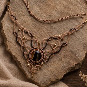 May include: A macrame necklace featuring a central round tiger eye gemstone surrounded by intricate tan and dark brown knotted cord patterns, accented with small metallic beads and a thin adjustable cord closure for a secure fit around the neck.