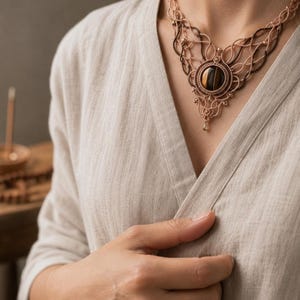 May include: A macrame necklace featuring a central round tiger eye gemstone set in intricate beige and dark brown knotted cord work. The design includes delicate woven patterns, small metallic beads, and a teardrop-shaped pendant accent.