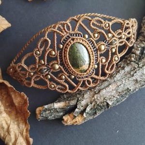 May include: A handcrafted macrame bracelet with an oval green stone at its centre. The bracelet is made with brown cord and small gold-coloured beads. The design has an open, intricate pattern. The bracelet is approximately 15 cm in length.