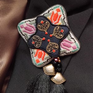 Puede incluir: Broche en forma de estrella con un centro azul marino con filigranas doradas y detalles rojos. El broche tiene un borde bordado colorido con motivos florales, borlas negras y dijes de metal dorado. Se muestra sobre un traje negro.