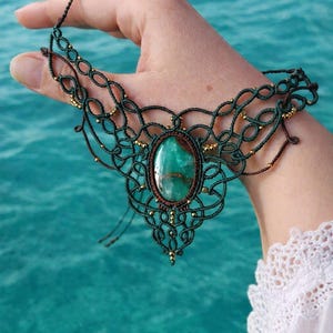 以下が含まれることがあります： 大きな楕円形の緑色の宝石をあしらった、手作りのマクラメネックレス。複雑な濃い緑色のマクラメのデザインは、小さな金のビーズでアクセントがつけられています。ネックレスはターコイズブルーの背景に展示されています。