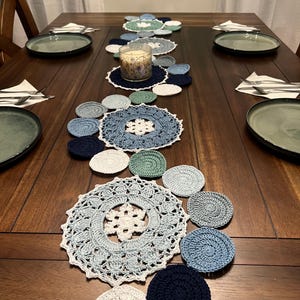 以下が含まれることがあります： 青、白、緑の色合いの手作りのかぎ針編みのドイリーとコースターがセンターピースとして配置された、食事用の木製ダイニングテーブル。 皿、カトラリー、キャンドルもテーブルにあります。