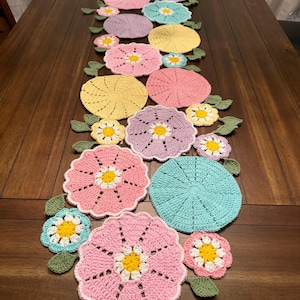 May include: A handmade, colorful crochet table runner featuring a floral design. The runner showcases various pastel-colored flowers, including pink, yellow, and blue, with green leaves. The flowers are arranged in a row, creating a decorative accent for a table.