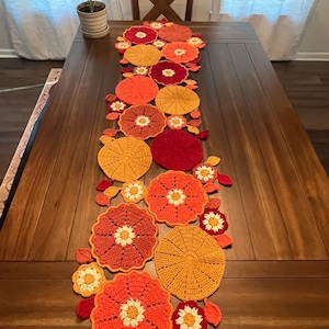 Può includere: Runner da tavolo all'uncinetto fatto a mano con un design floreale nei toni dell'arancione, del giallo e del rosso. Il runner è posto su un tavolo di legno scuro. Il motivo floreale comprende grandi fiori circolari e fiori di accento più piccoli.