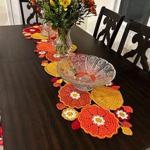 May include: A dark wooden dining table is decorated with a colourful, handmade crochet table runner. The runner features orange, yellow, and red floral designs. A clear glass bowl and a vase of flowers complete the table setting.
