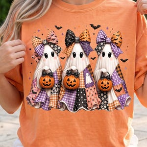 May include: An orange t-shirt featuring a graphic of three white ghosts wearing large patterned bows. Each ghost holds a small orange jack-o'-lantern bucket. The ghosts are decorated with various plaid and checkered patterns, surrounded by small black bats.