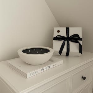 May include: A white, bowl-shaped candle with several wicks sits on a white book with the word "WHITE" printed on it. A white gift box tied with a black ribbon is also present. The items are on a white chest of drawers.
