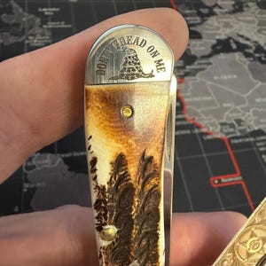 May include: A folding pocket knife with a handle featuring a forest scene and a metal top with the words "DON'T TREAD ON ME" and a coiled snake graphic. The knife has a gold and brown colour scheme.