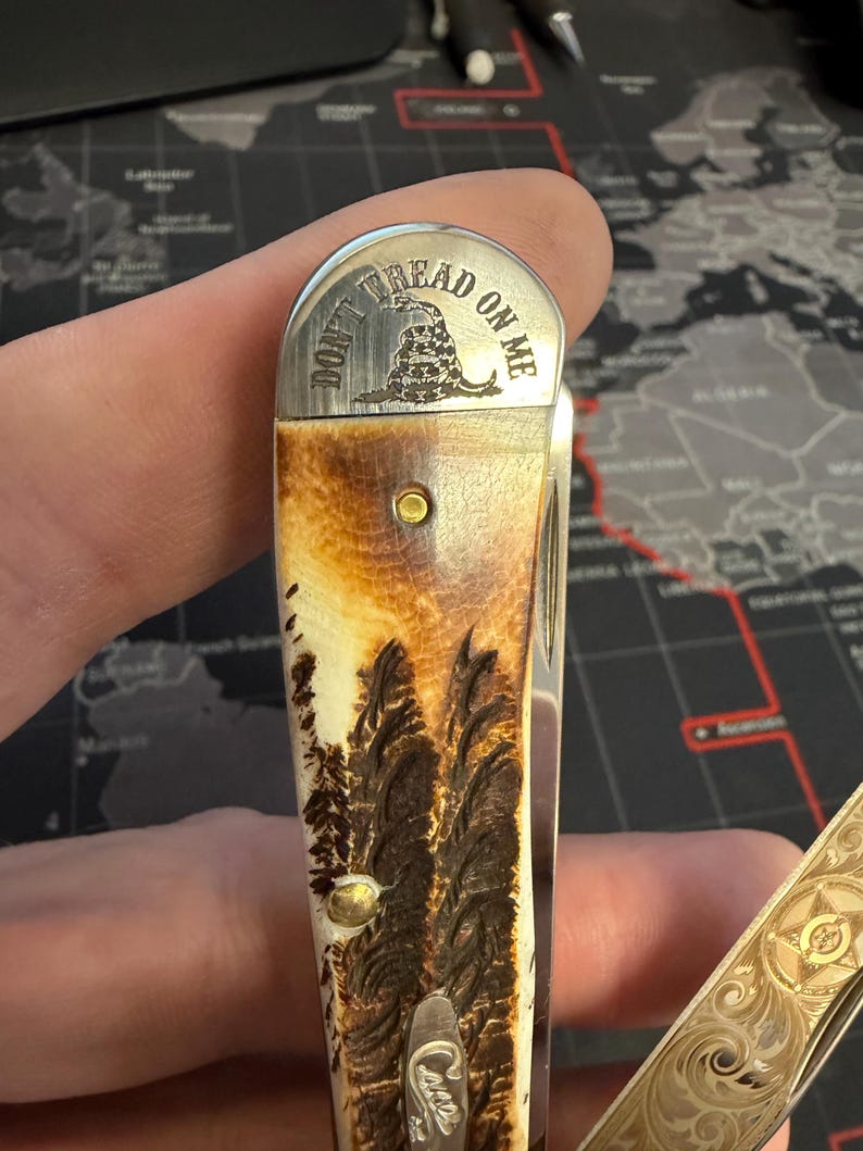 May include: A folding pocket knife with a handle featuring a forest scene and a metal top with the words "DON'T TREAD ON ME" and a coiled snake graphic. The knife has a gold and brown colour scheme.