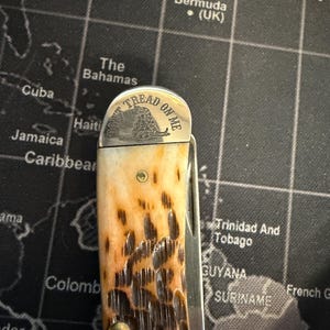 May include: A pocket knife with a patterned brown and tan handle. The blade is closed. The top of the knife has the words "DON'T TREAD ON ME" and a coiled snake.