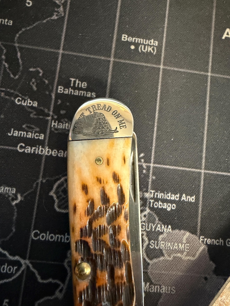 May include: A pocket knife with a patterned brown and tan handle. The blade is closed. The top of the knife has the words "DON'T TREAD ON ME" and a coiled snake.