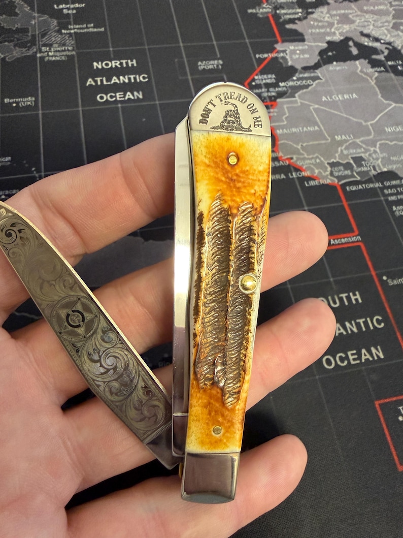 May include: A folding pocket knife with a cream and brown handle, featuring a detailed textured design. The blade is open, revealing an engraved pattern. The knife's handle has the text "DON'T TREAD ON ME" and a coiled snake.