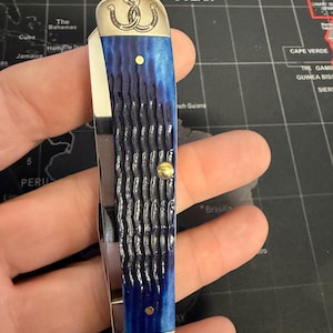 May include: A pocket knife with a blue handle and a silver blade. The handle has a textured pattern and is accented with gold-colored rivets. The top of the knife features a silver emblem with two intertwined horseshoes.