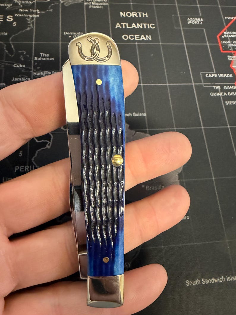 May include: A pocket knife with a blue handle and a silver blade. The handle has a textured pattern and is accented with gold-colored rivets. The top of the knife features a silver emblem with two intertwined horseshoes.