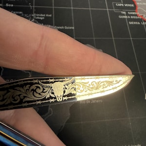 May include: A small, decorative knife with a gold-colored blade and a black handle. The blade features an engraved bull skull and floral design with barbed wire accents. The knife is held against a world map background.