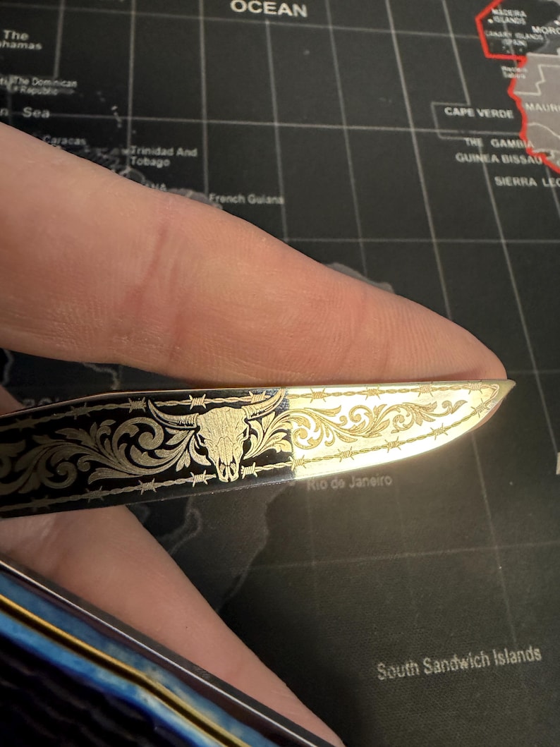 May include: A small, decorative knife with a gold-colored blade and a black handle. The blade features an engraved bull skull and floral design with barbed wire accents. The knife is held against a world map background.