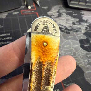 May include: A folding pocket knife with a brown and cream-coloured patterned handle. The metal top of the knife is engraved with the words "DON'T TREAD ON ME" and a coiled snake image.