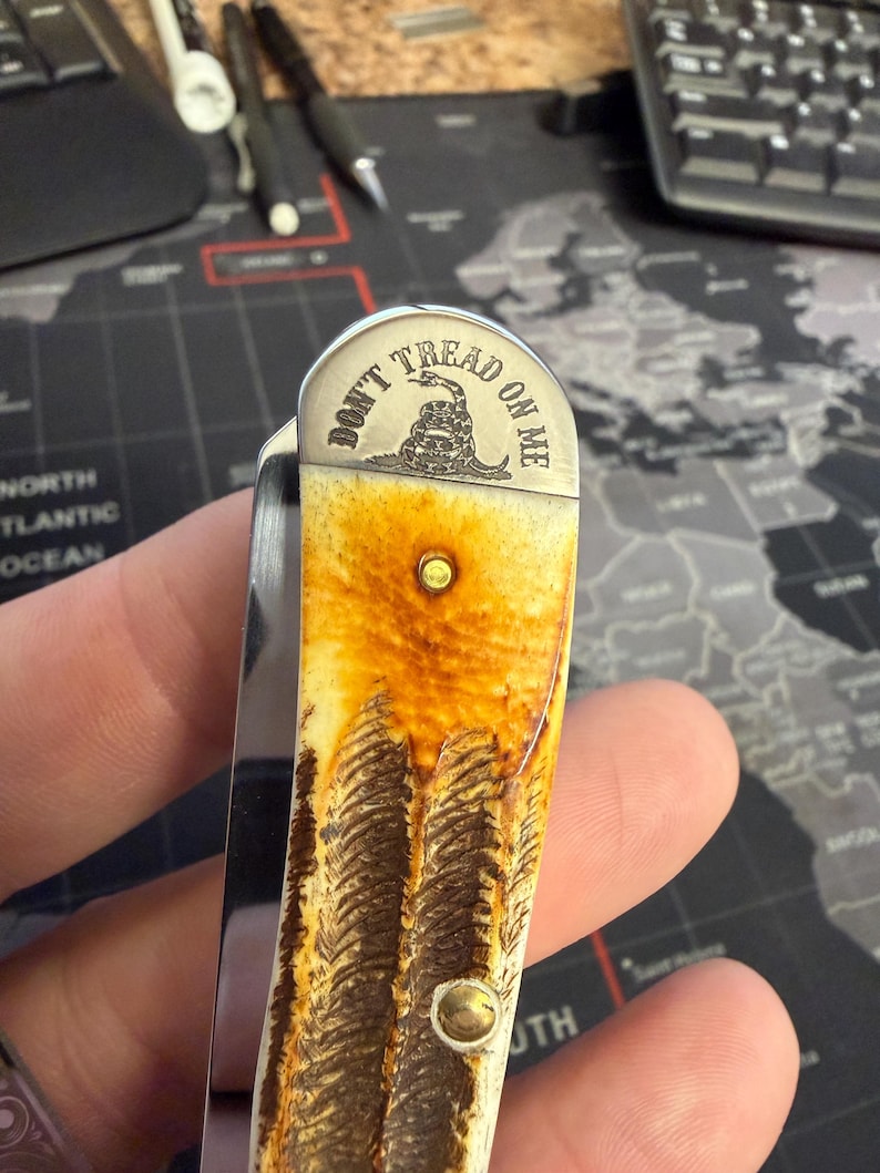 May include: A folding pocket knife with a brown and cream-coloured patterned handle. The metal top of the knife is engraved with the words "DON'T TREAD ON ME" and a coiled snake image.