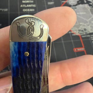 May include: Pocket knife with a blue and black handle and a silver blade. The knife features a decorative silver top with a horseshoe design. The handle has two gold-colored rivets.