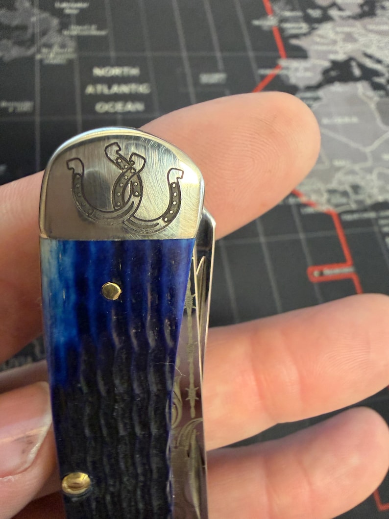 May include: Pocket knife with a blue and black handle and a silver blade. The knife features a decorative silver top with a horseshoe design. The handle has two gold-colored rivets.