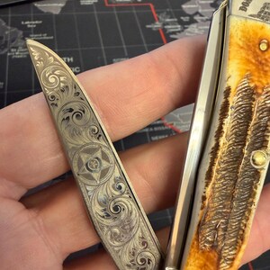 May include: A pocket knife with an engraved blade, featuring a star and scroll pattern. The handle has a textured, light brown appearance. The knife is open, displaying the detailed engraving on the blade.