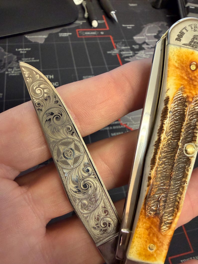 May include: A pocket knife with an engraved blade, featuring a star and scroll pattern. The handle has a textured, light brown appearance. The knife is open, displaying the detailed engraving on the blade.