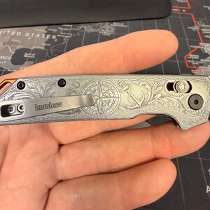 May include: A silver folding knife with an engraved nautical design featuring a compass rose and anchor. The knife has a pocket clip and the brand name "Kershaw" is visible. The blade is closed, and the knife is held in a hand.