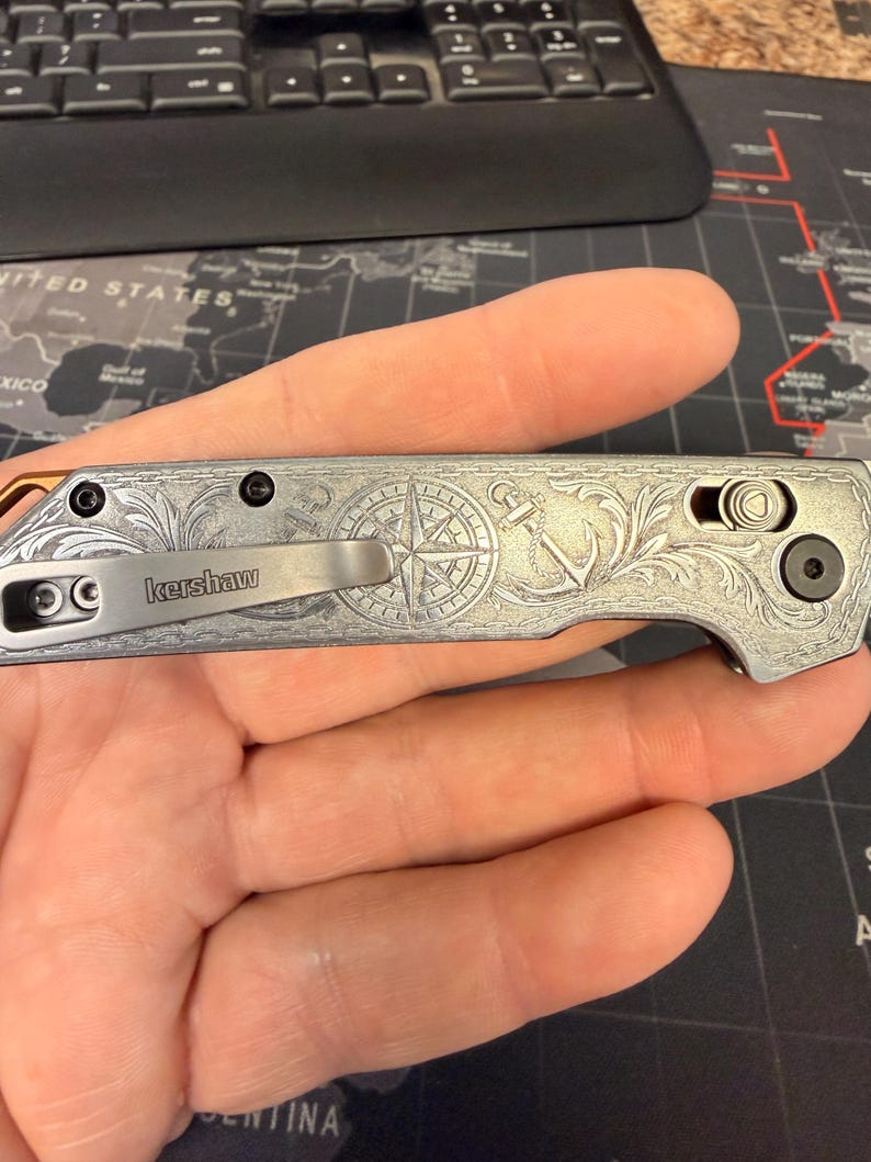 May include: A silver folding knife with an engraved nautical design featuring a compass rose and anchor. The knife has a pocket clip and the brand name "Kershaw" is visible. The blade is closed, and the knife is held in a hand.