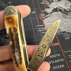 May include: A folding pocket knife with an engraved blade and handle. The handle has a brown and cream pattern with a silver emblem. The open blade displays detailed floral designs. The text "DON'T TREAD ON ME" is visible on the handle.