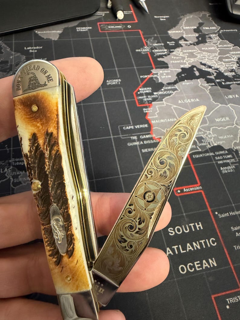 May include: A folding pocket knife with an engraved blade and handle. The handle has a brown and cream pattern with a silver emblem. The open blade displays detailed floral designs. The text "DON'T TREAD ON ME" is visible on the handle.