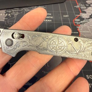 May include: A silver folding knife with intricate engraved nautical designs. The knife features a compass rose, anchors, and floral patterns. The blade is closed, and the handle is held in a hand, showcasing the detailed craftsmanship.